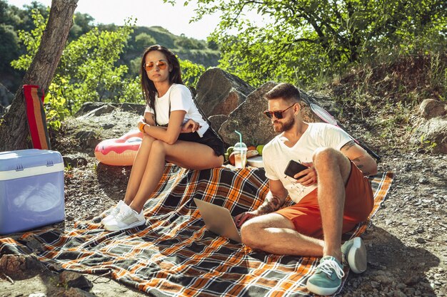 Pareja joven haciendo un picnic en la orilla del río en un día soleado. Mujer y hombre pasando tiempo juntos en la naturaleza. Divertirse, comer, jugar y reír. Concepto de relación, amor, verano, fin de semana.