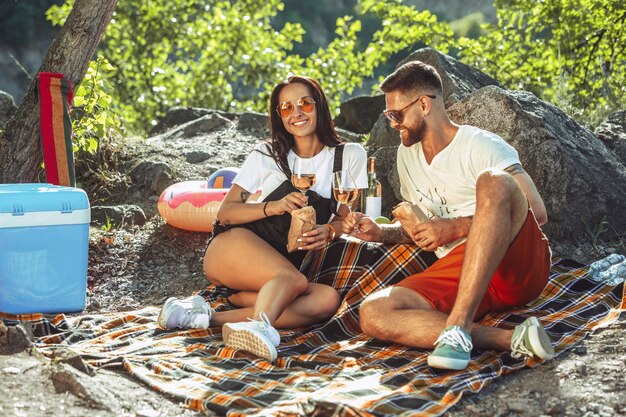 Pareja joven haciendo un picnic en la orilla del río en un día soleado. Mujer y hombre pasando tiempo juntos en la naturaleza. Divertirse, comer, jugar y reír. Concepto de relación, amor, verano, fin de semana.