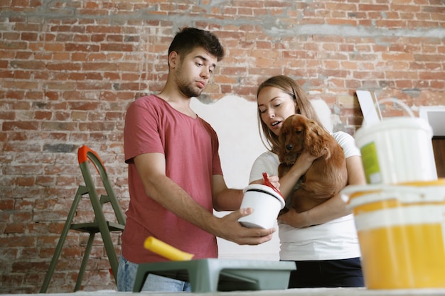 Pareja joven haciendo reparación de apartamentos juntos ellos mismos. Hombre y mujer casados haciendo remodelación o renovación de la casa. Concepto de relaciones, familia, mascota, amor. Elegir el color de la pintura, sujetando al perro.