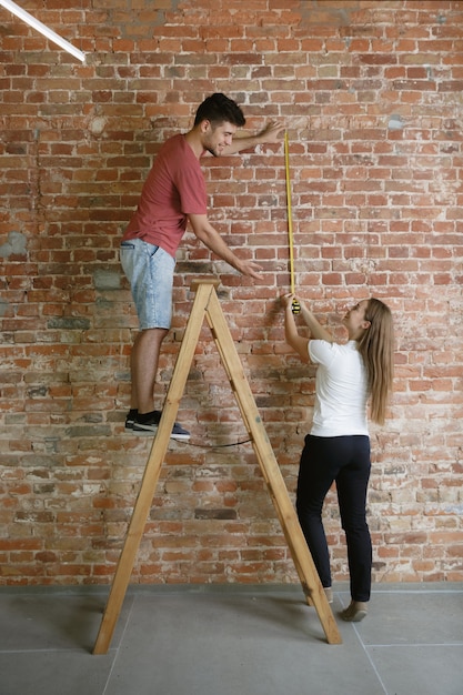 Pareja joven haciendo reparación de apartamentos juntos ellos mismos. Hombre y mujer casados haciendo remodelación o renovación de la casa. Concepto de relaciones, familia, mascota, amor. Medición de pie en escalera con el metro.