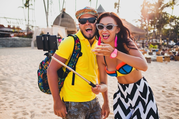 Pareja joven hermosa hipster enamorada en la playa tropical, tomando fotos selfie en teléfono inteligente, gafas de sol, atuendo elegante, vacaciones de verano, divirtiéndose, sonriendo, feliz, colorido, emoción positiva