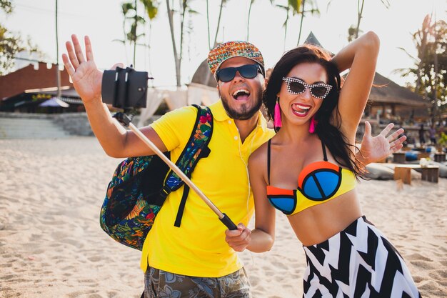 Pareja joven hermosa hipster enamorada en la playa tropical, tomando fotos selfie en teléfono inteligente, gafas de sol, atuendo elegante, vacaciones de verano, divirtiéndose, sonriendo, feliz, colorido, emoción positiva