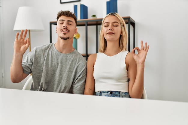 Foto gratuita una pareja joven y hermosa con ropa informal sentada en la mesa en casa se relaja y sonríe con los ojos cerrados haciendo gestos de meditación con los dedos. concepto de yoga