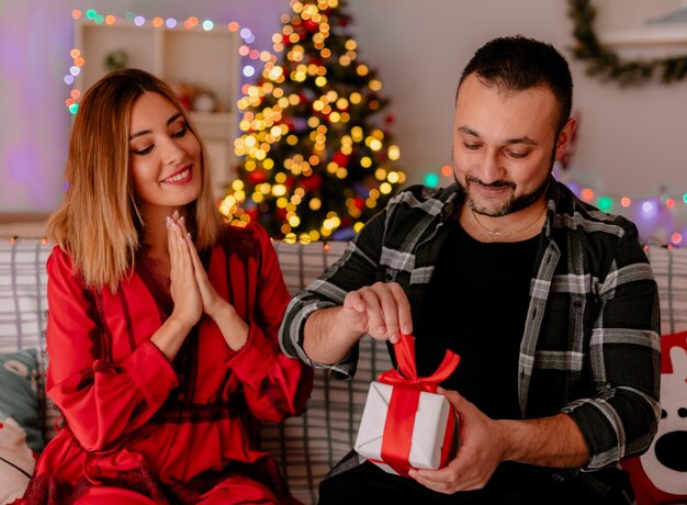 Pareja joven y hermosa sentada en un sofá hombre abriendo un regalo mientras su novia feliz mirándolo celebrando la Navidad juntos en una habitación decorada con árbol de Navidad en el fondo