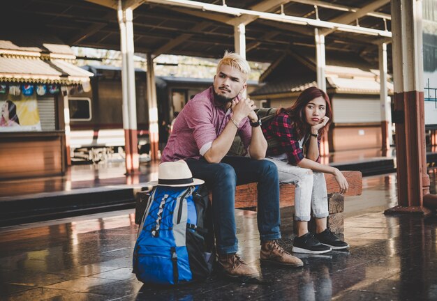 Pareja joven hipster sentado en el banco de madera en la estación de tren. Par sentado esperando el tren en la plataforma.