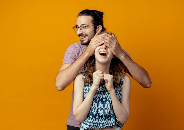 Pareja joven hombre cerrando los ojos de la niña mientras ella sonríe feliz y emocionada sobre la pared naranja