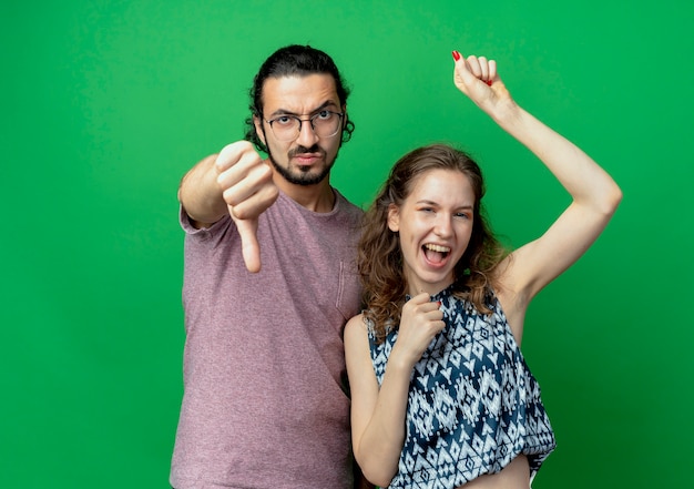 Pareja joven hombre disgustado mostrando los pulgares hacia abajo de pie junto a su novia feliz sobre la pared verde