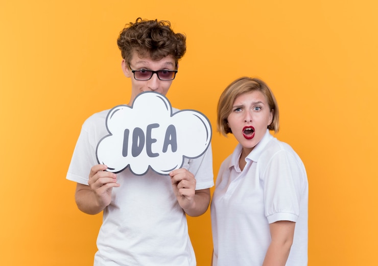 Foto gratuito pareja joven hombre feliz con cartel de burbujas de discurso con idea de palabra mientras su novia confundida de pie sobre la pared naranja
