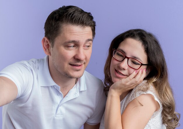 Pareja joven hombre feliz mirando a su amada novia sonriente de pie sobre la pared azul