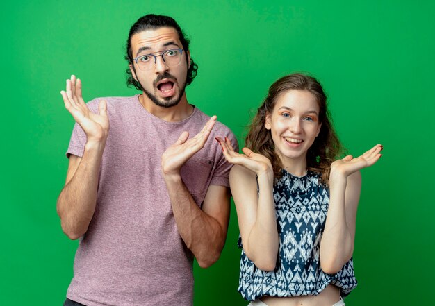 Pareja joven hombre y mujer, confundidos extendiendo los brazos hacia los lados de pie sobre la pared verde