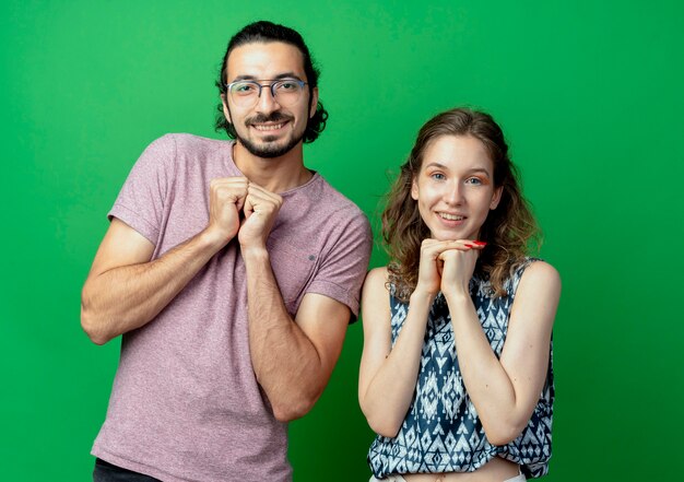 Pareja joven hombre y mujer, felices y positivos tomados de la mano juntos esperando sorpresa de pie sobre la pared verde