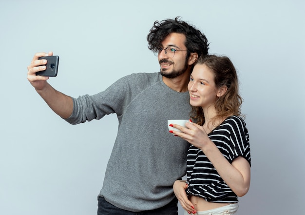 Pareja joven hombre y mujer, hombre feliz tomando una foto de ellos con su teléfono inteligente mientras su novia de pie junto a él tomando café sobre una pared blanca