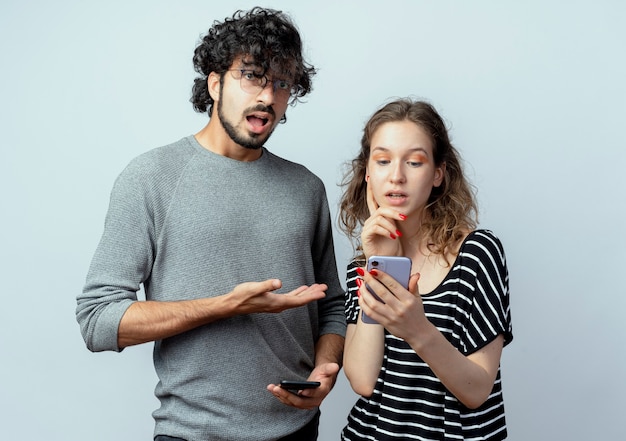 Pareja joven hombre y mujer, hombre molesto apuntando a su novia que sostiene smartphone de pie sobre fondo blanco.