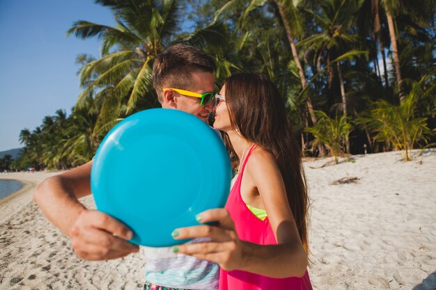 Pareja joven hombre y mujer jugando disco volador en playa tropical, vacaciones de verano, amor, romance, estado de ánimo feliz, sonriendo, divirtiéndose, atuendo hipster, gafas de sol, pantalones cortos de mezclilla, estado de ánimo soleado y positivo