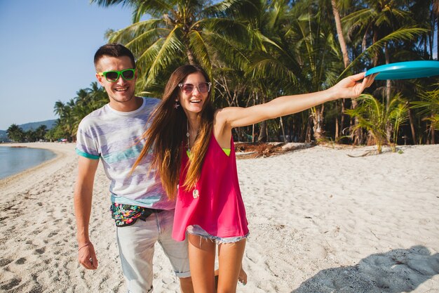 Pareja joven hombre y mujer jugando disco volador en playa tropical, vacaciones de verano, amor, romance, estado de ánimo feliz, sonriendo, divirtiéndose, atuendo hipster, gafas de sol, pantalones cortos de mezclilla, estado de ánimo soleado y positivo