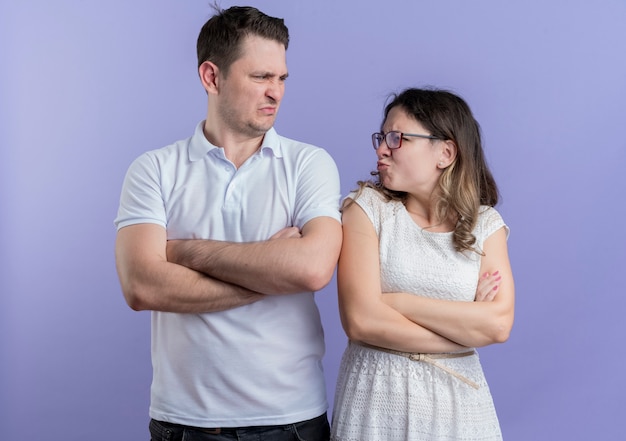 Pareja joven hombre y mujer mirando el uno al otro frunciendo el ceño con los brazos cruzados de pie sobre la pared azul