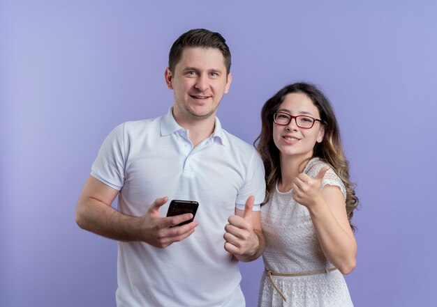 Pareja joven hombre y mujer mirando a la cámara sosteniendo el teléfono inteligente mostrando los pulgares para arriba de pie sobre la pared azul