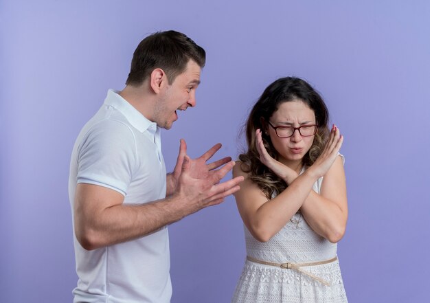 Pareja joven hombre y mujer peleando hombre gritando a su novia mientras ella hace la señal de pare cruzando las manos de pie sobre la pared azul