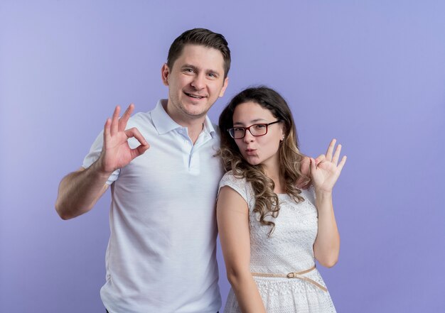 Pareja joven hombre y mujer de pie juntos mostrando ok canta sonriendo sobre la pared azul