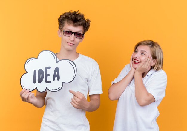 Pareja joven hombre y mujer de pie juntos sonriendo feliz y positivo sosteniendo el letrero de burbujas de discurso con idea de palabra sobre pared naranja