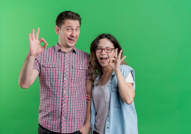 Pareja joven hombre y mujer en ropa casual feliz y positivo mostrando signos de ok divirtiéndose juntos de pie sobre la pared verde