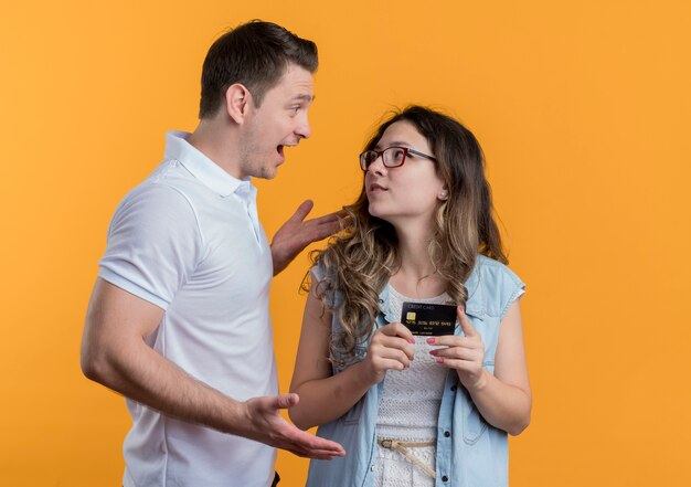 Pareja joven hombre y mujer en ropa casual hombre hablando con su novia sonriente de pie sobre la pared naranja