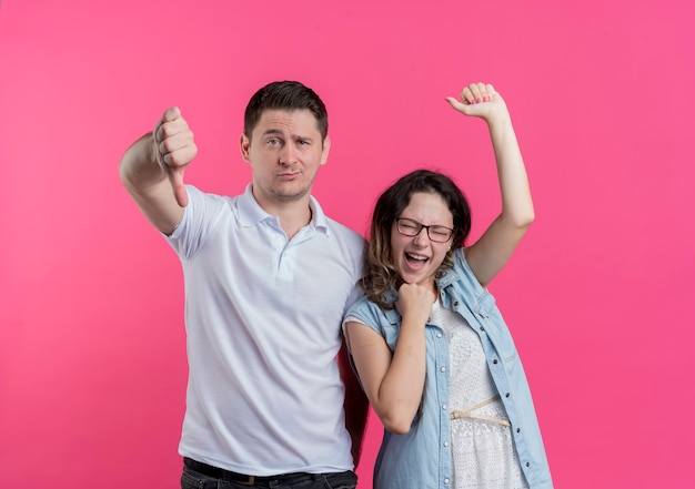 Pareja joven hombre y mujer en ropa casual hombre mostrando los pulgares hacia abajo mientras su novia feliz y regocijo de pie sobre la pared rosa