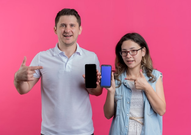 Foto gratuita pareja joven hombre y mujer en ropa casual mostrando smartphones apuntando con los dedos a ellos sonriendo sobre rosa