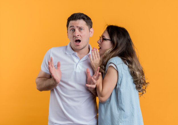 Pareja joven hombre y mujer en ropa casual mujer susurrando un secreto a su novio sorprendido de pie sobre la pared naranja