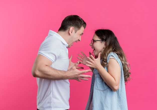 Pareja joven hombre y mujer en ropa casual peleando gritando el uno al otro de pie sobre la pared rosa