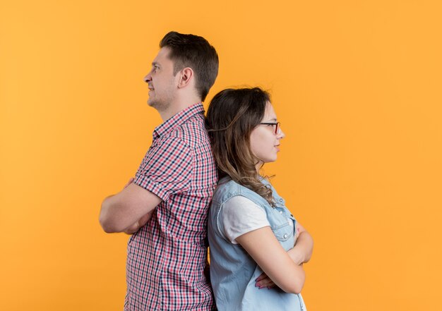 Pareja joven hombre y mujer en ropa casual de pie espalda con espalda después de la pelea de pie sobre la pared naranja