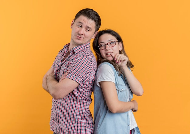 Foto gratuita pareja joven hombre y mujer en ropa casual de pie espalda con espalda sonriendo sobre pared naranja