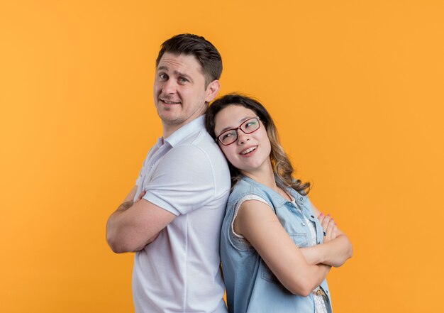 Pareja joven hombre y mujer en ropa casual de pie juntos espalda con espalda feliz y positivo sonriendo sobre pared naranja