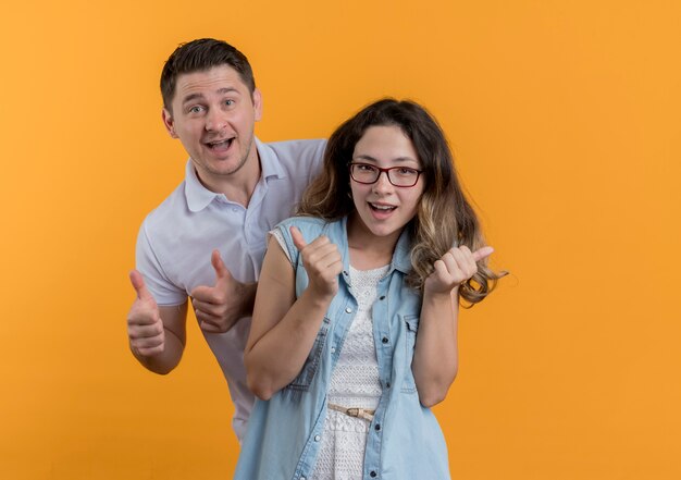 Pareja joven hombre y mujer en ropa casual sonriendo alegremente feliz y emocionado mostrando los pulgares para arriba de pie sobre la pared naranja