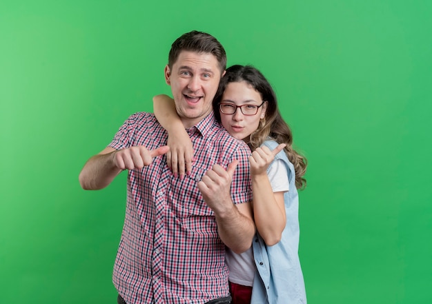 Pareja joven hombre y mujer en ropa casual sonriendo mostrando los pulgares para arriba de pie sobre la pared verde