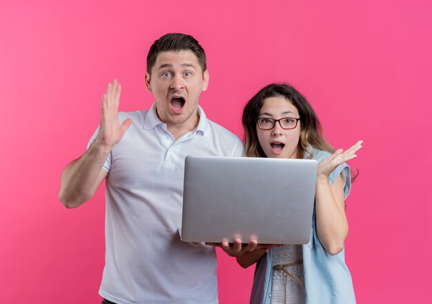 Pareja joven hombre y mujer en ropa casual sosteniendo portátil mirando asombrado y sorprendido de pie sobre la pared rosa