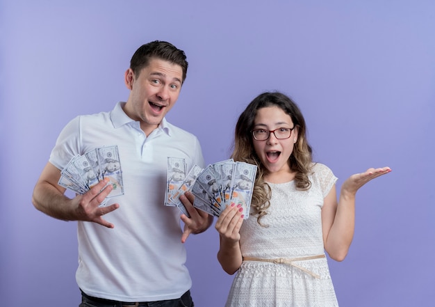 Foto gratuita pareja joven, hombre y mujer, tenencia, efectivo, feliz y sorprendido, posición, juntos, encima, pared azul