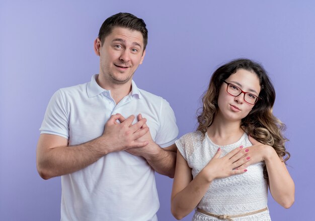 Pareja joven hombre y mujer tomados de la mano en el pecho sintiéndose agradecido, feliz y positivo de pie sobre la pared azul