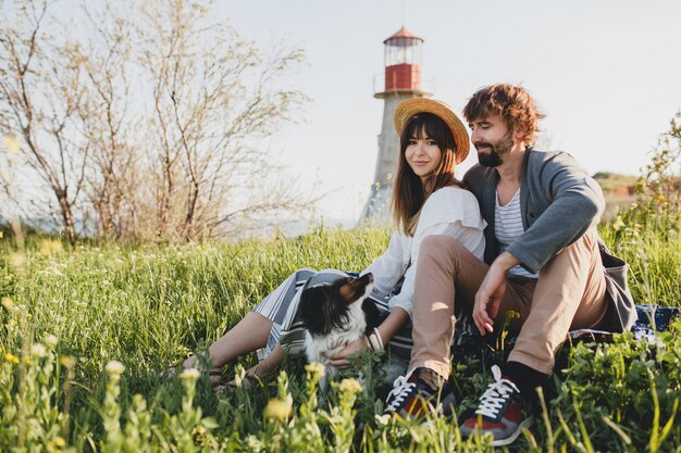 Pareja joven inconformista con estilo en el amor con el perro en el campo, sentado en la hierba