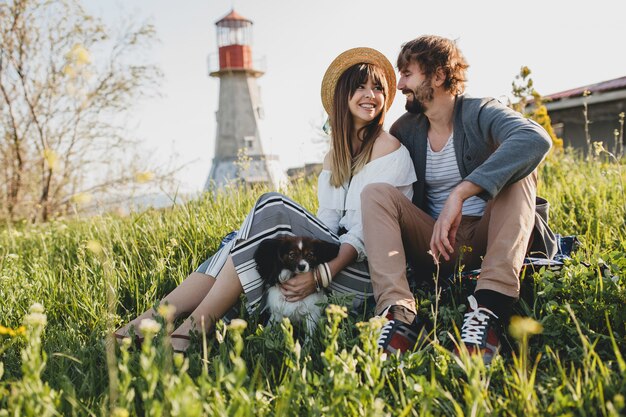 Pareja joven inconformista con estilo en el amor con el perro en el campo, sentado en la hierba