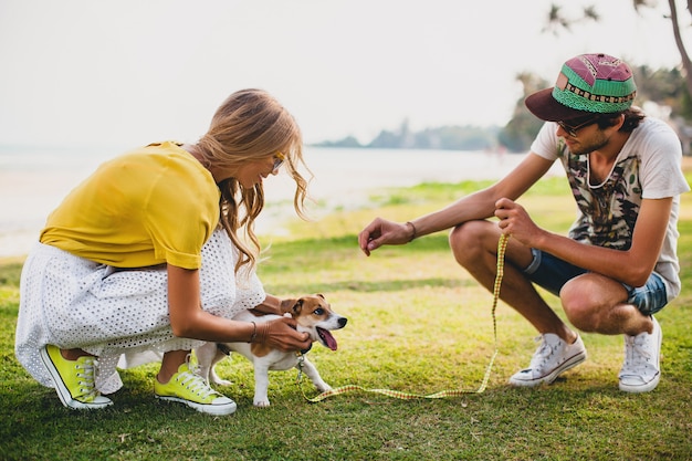 Pareja joven inconformista con estilo enamorado caminando y jugando con perro en playa tropical