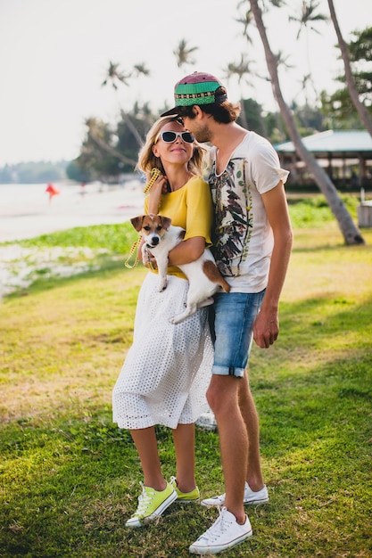 Pareja joven inconformista con estilo enamorado sosteniendo un perro en el parque tropical, sonriendo y divirtiéndose durante sus vacaciones