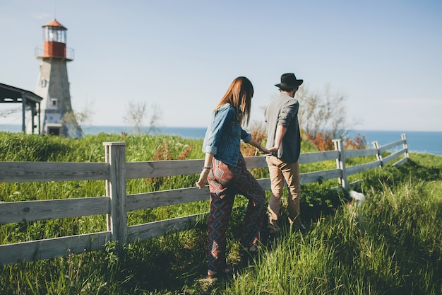 Pareja joven inconformista estilo indie enamorado caminando en el campo, tomados de la mano, faro en el fondo, cálido día de verano, soleado, traje bohemio, sombrero