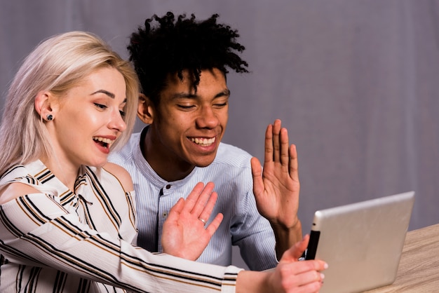 Pareja joven interracial haciendo videollamada en tableta digital agitando sus manos