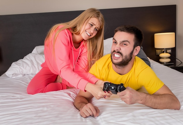 Pareja joven jugando videojuegos en la cama. Chico jugando un videojuego, ella se queja de que no le prestó atención