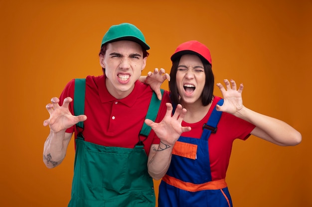 Pareja joven juguetona en uniforme de trabajador de la construcción y gorra haciendo gesto de patas de tigre chico mirando a la cámara chica con los ojos cerrados aislado en la pared naranja