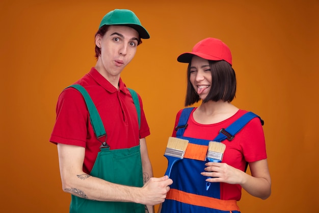 Pareja joven juguetona en uniforme de trabajador de la construcción y gorra sosteniendo pinceles delante del pecho de las niñas chico sorprendido chica tonta mostrando la lengua con los ojos cerrados aislados