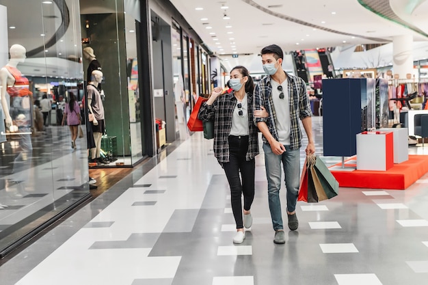 Pareja joven en máscara de protección sosteniendo múltiples bolsas de papel caminando en el pasillo del gran centro comercial