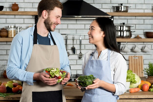 Pareja joven mirando el uno al otro mientras sostiene un tazón de ensalada y hojas verdes frescas