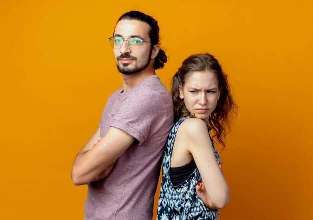 Pareja joven mirando a la cámara de pie espalda con espalda con los brazos cruzados sobre fondo naranja